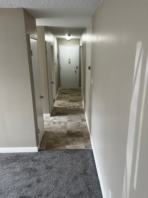 Long hallway with white walls, multiple closed doors on both sides, and mixed flooring of carpet and tile under bright overhead lighting.
