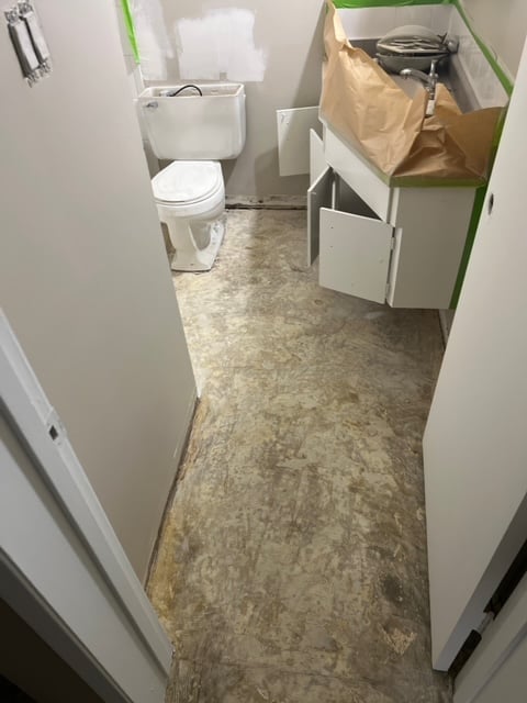 Narrow bathroom under renovation with white toilet, exposed pipes along green-taped walls, and brown paper-covered fixtures on the right side