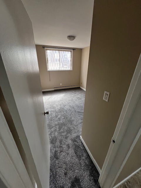 Interior hallway view of empty apartment room with gray carpeting, beige walls, window with blinds, and ceiling smoke detector
